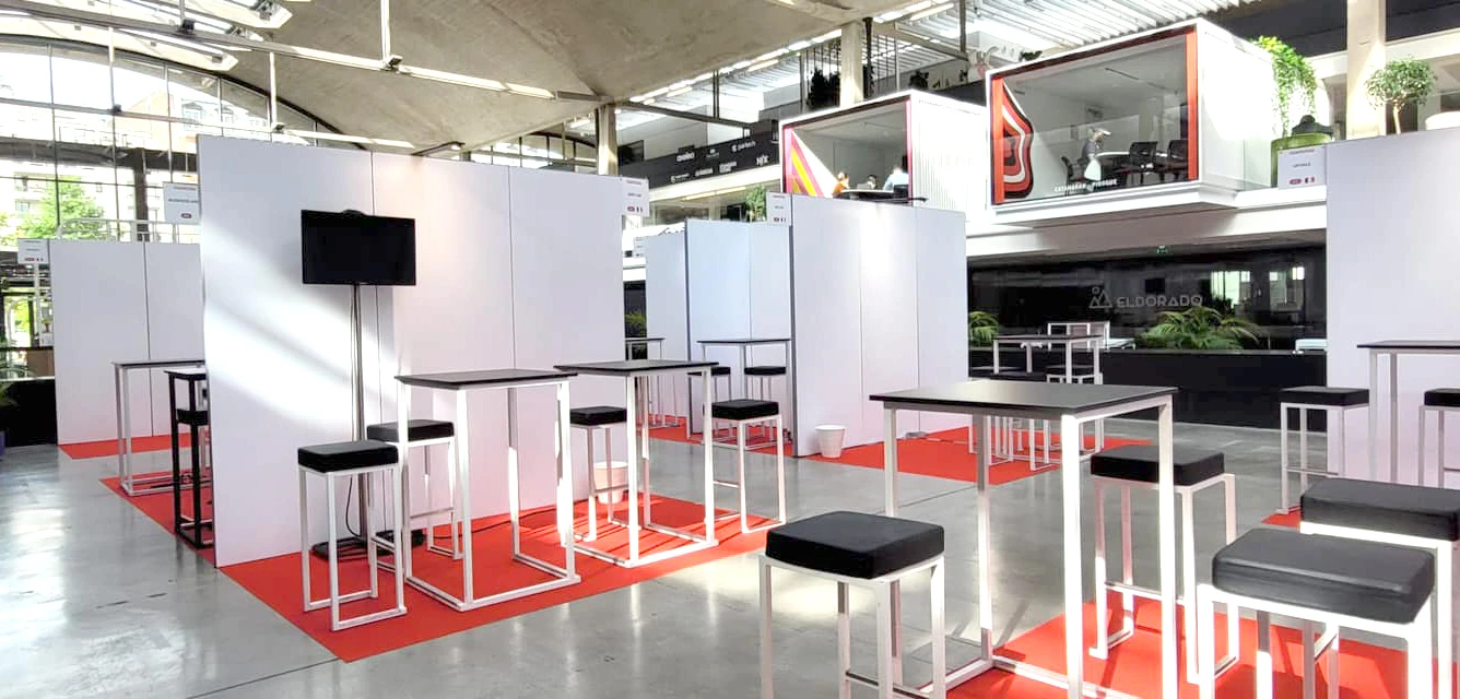 Espace networking sur moquette rouge avec mange-debout blancs à plateau noir, tabourets hauts noirs et cloisons blanches dans un hall vitré
